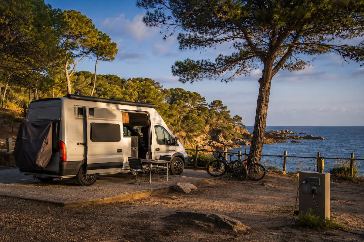 Área para autocaravanas junto al mar en la Costa Brava, con camper, bicicletas, punto de luz y vistas a una costa rocosa al atardecer.